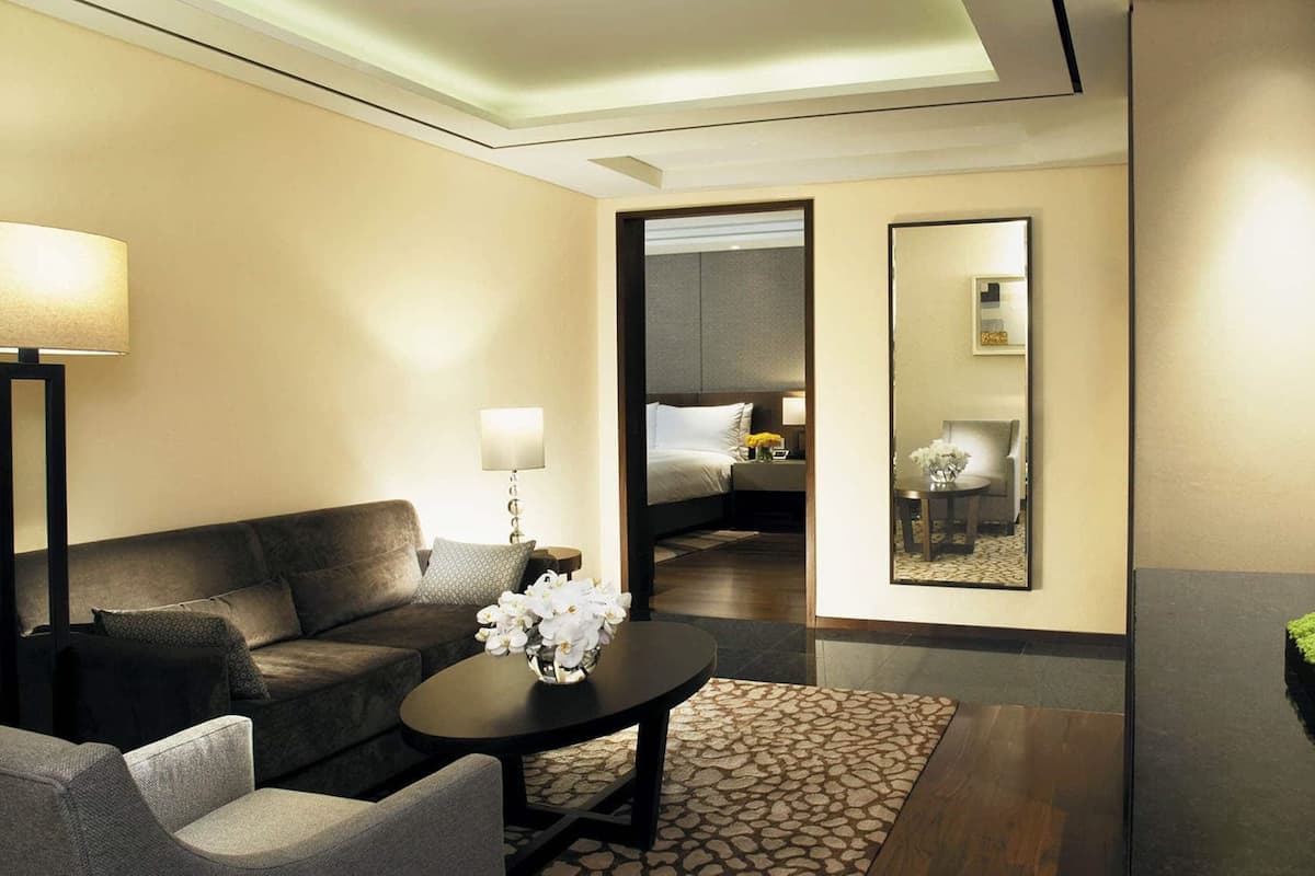 A modern hotel room with a dark-colored sofa, a small round table with a vase of white flowers, a large mirror, and a flat-screen TV mounted on the wall.