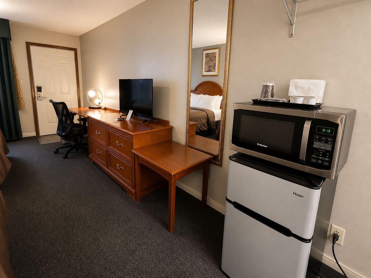A hotel room with a wooden desk, a television, a mirror, a bed, and a refrigerator.