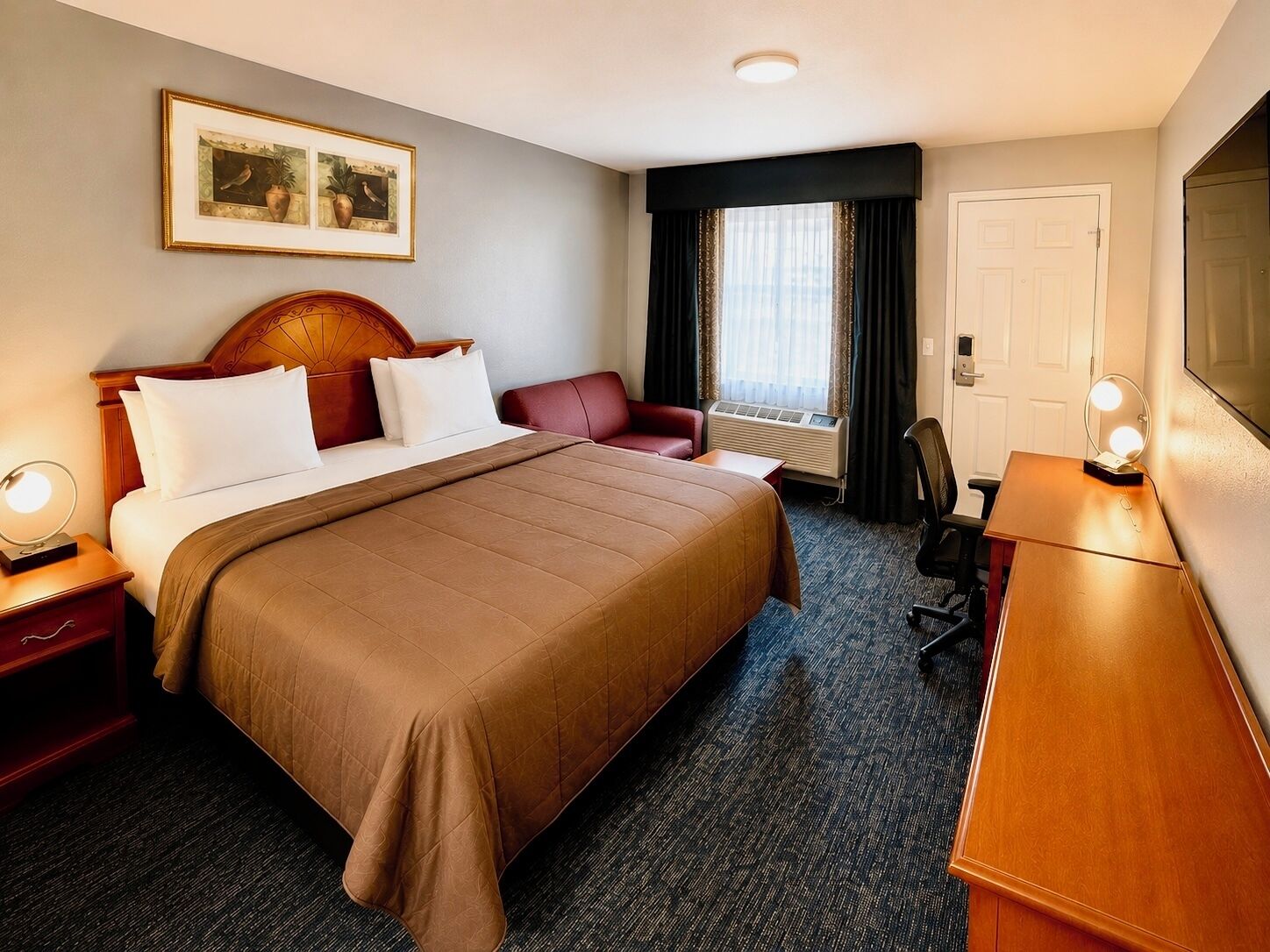 A hotel room with a large bed, a desk with a mirror, a chair, a bedside table, a window with curtains, and a framed picture on the wall.