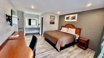 A hotel room with a bed, a desk, a chair, a small bedside table, a mirror, and a picture on the wall.