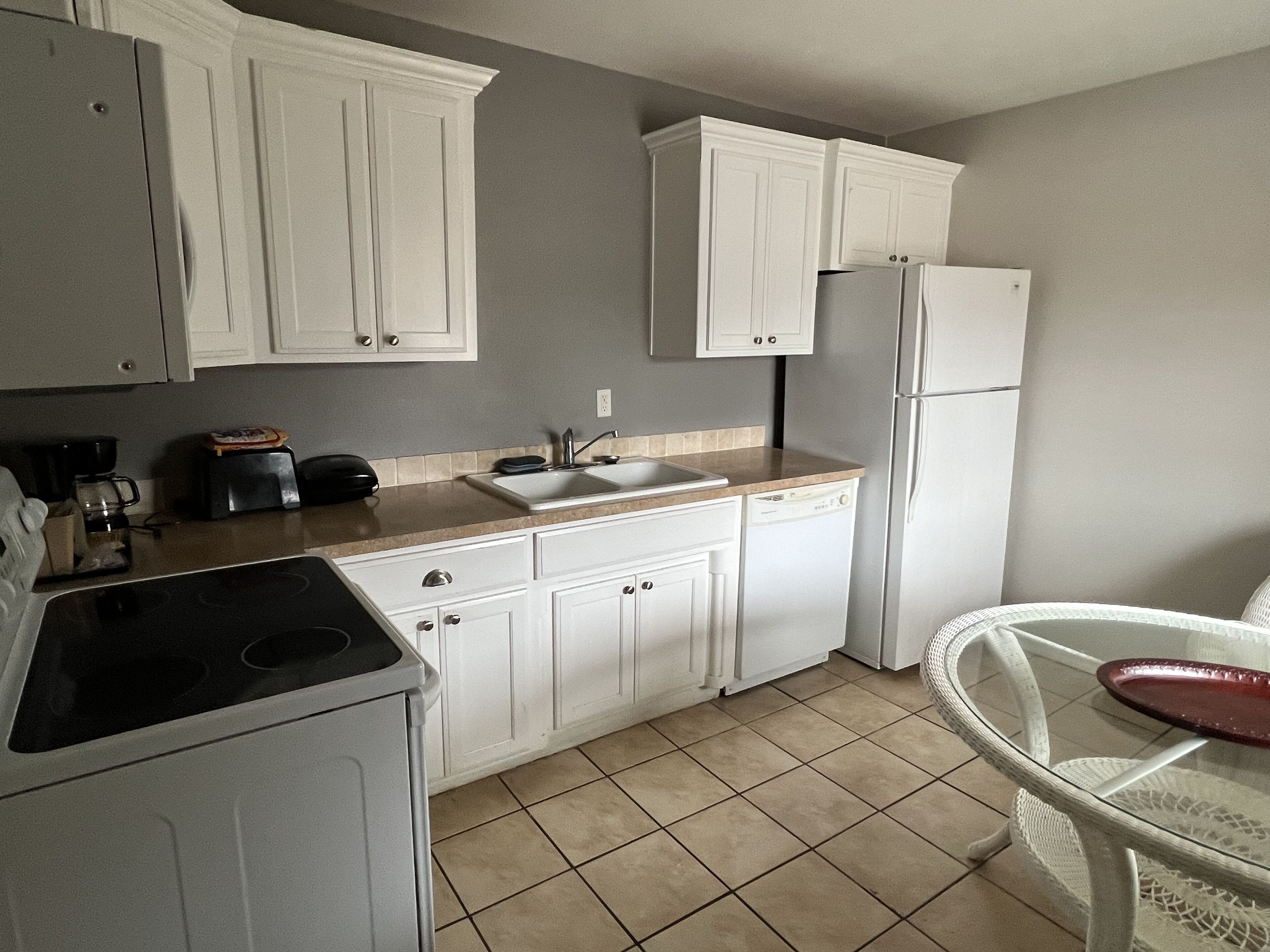 Cocina compacta con gabinetes blancos, lavavajillas, refrigerador, estufa y una pequeña mesa de comedor con una silla.
