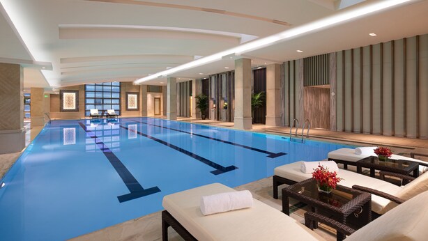 Ein großer, überdachter Pool mit Liegestühlen und Tischen, umgeben von Wänden mit vertikalen Linien und einer hohen Decke mit eingelassener Beleuchtung.