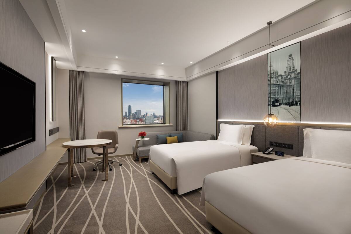 Habitación de hotel con dos camas, un escritorio, una silla, una mesita con un florero, un ventanal con vistas a la ciudad y un televisor montado.