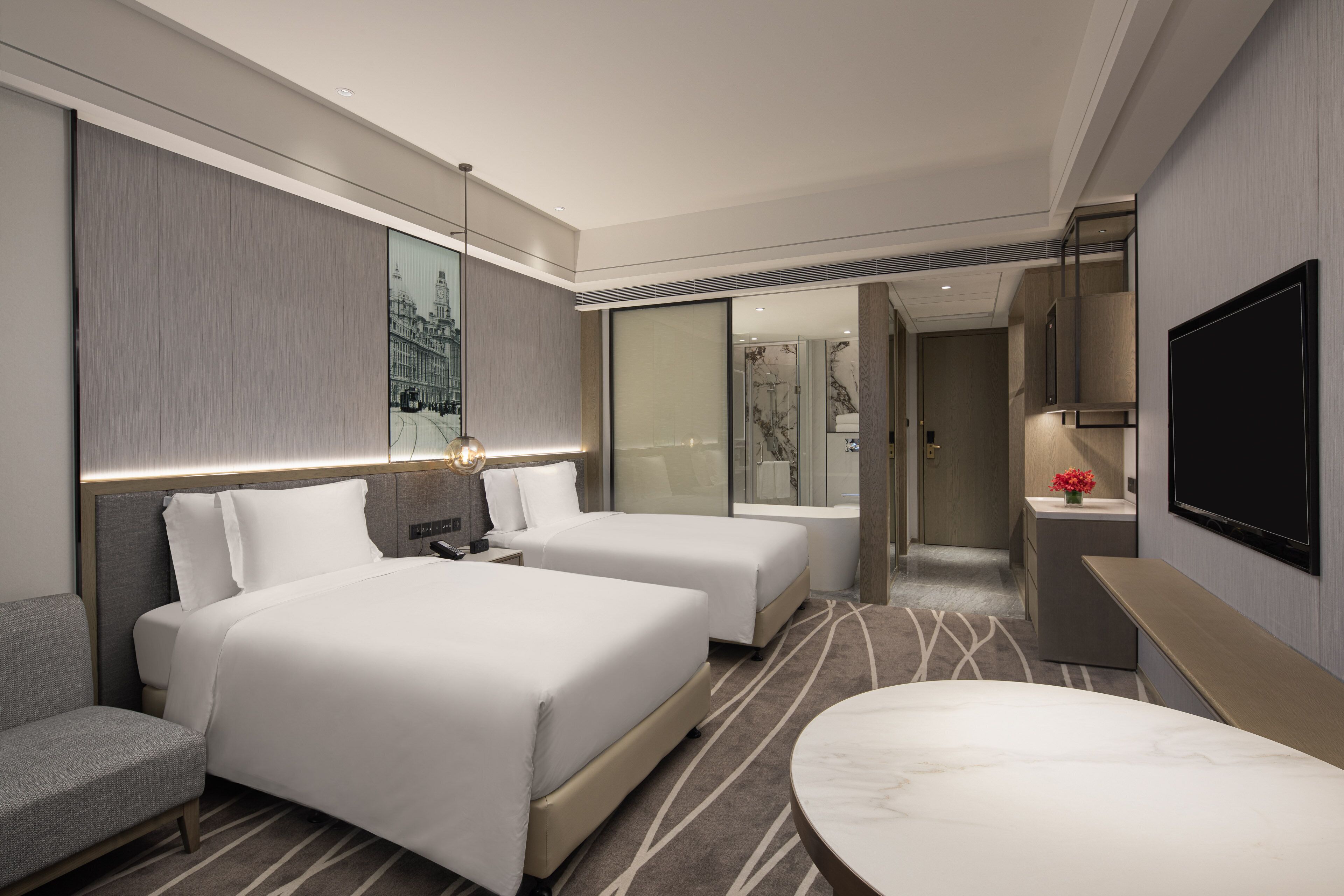 A hotel room with two beds, a sofa, a marble-topped table, a TV, and a bathroom visible through a glass door.