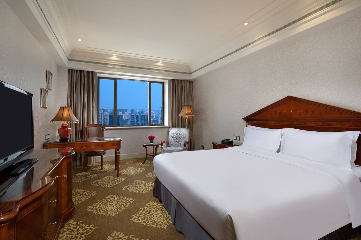 Habitación de hotel con una cama grande, un escritorio, una silla, un televisor y vistas a la ciudad a través de la ventana.