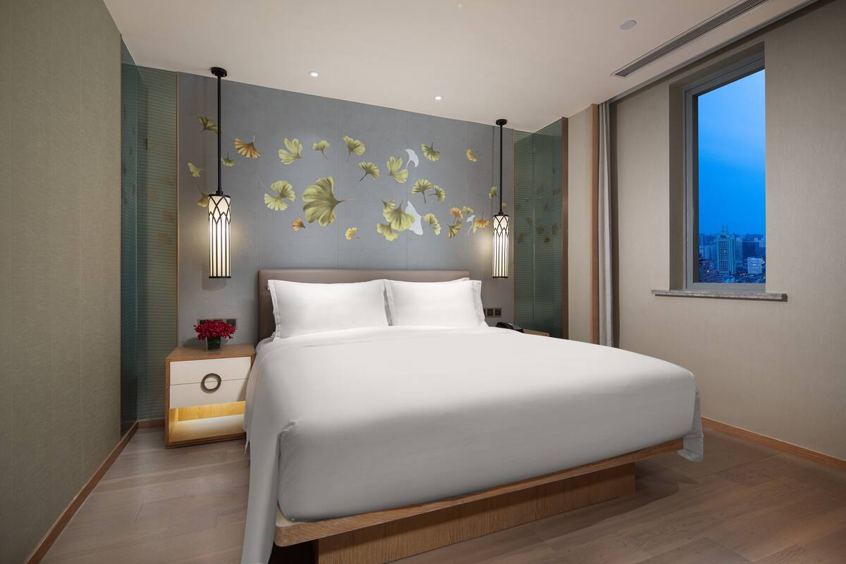 Un dormitorio moderno con una cama grande, dos mesitas de noche, una mesita auxiliar, una pared decorativa con motivos de hojas y vistas a la ciudad a través de un ventanal amplio.