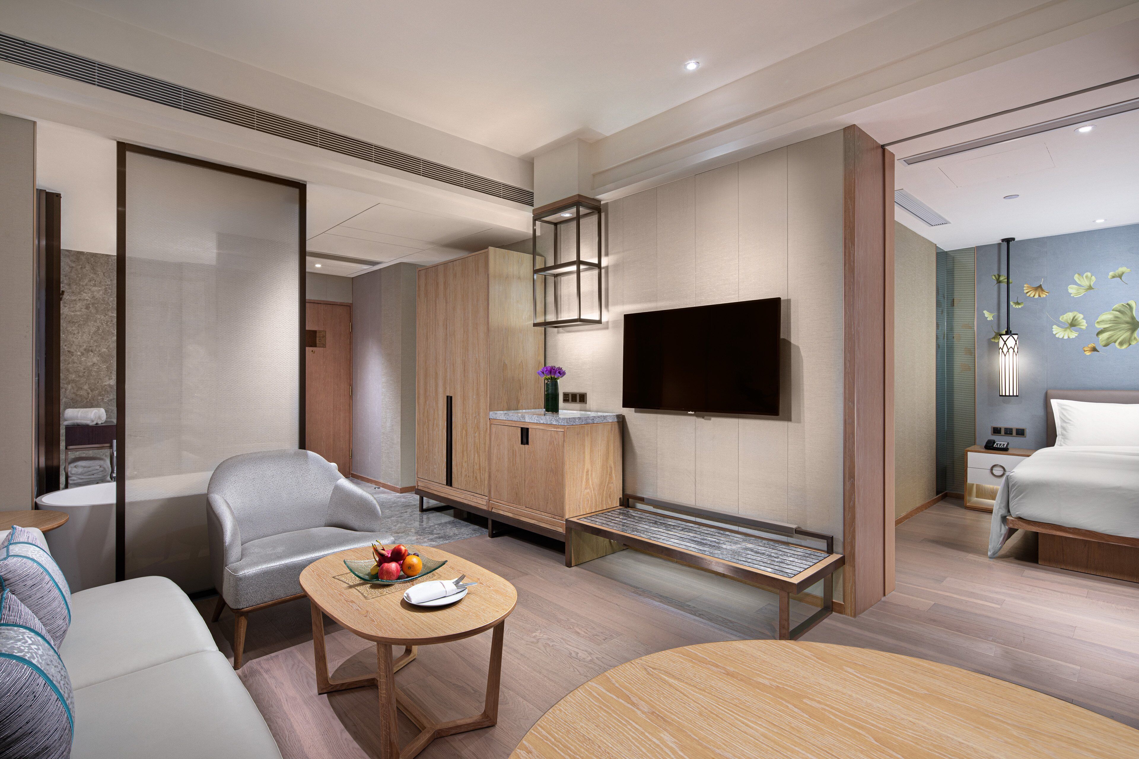 A modern hotel room with a sofa, a dining table with fruits, a TV, and a bed.