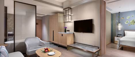 A modern hotel room with a sofa, a dining table with fruits, a TV, and a bed.
