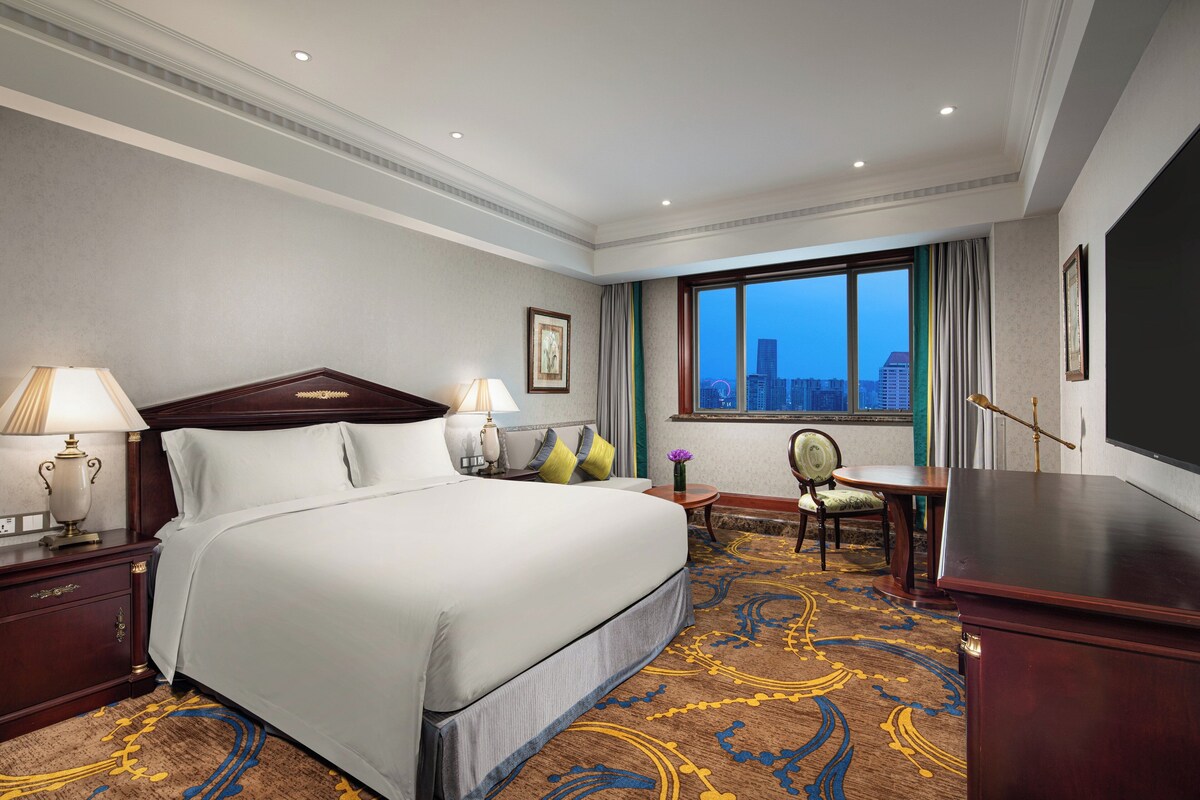 Une chambre d’hôtel avec un grand lit, un bureau, une chaise et une vue sur le paysage urbain par la fenêtre.