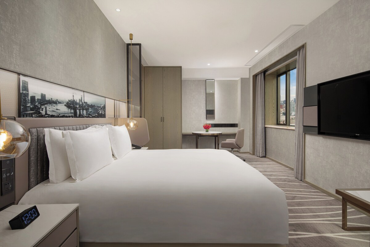 Une chambre d’hôtel moderne avec un grand lit, un bureau, une télévision et un tableau représentant un paysage urbain accroché au mur.