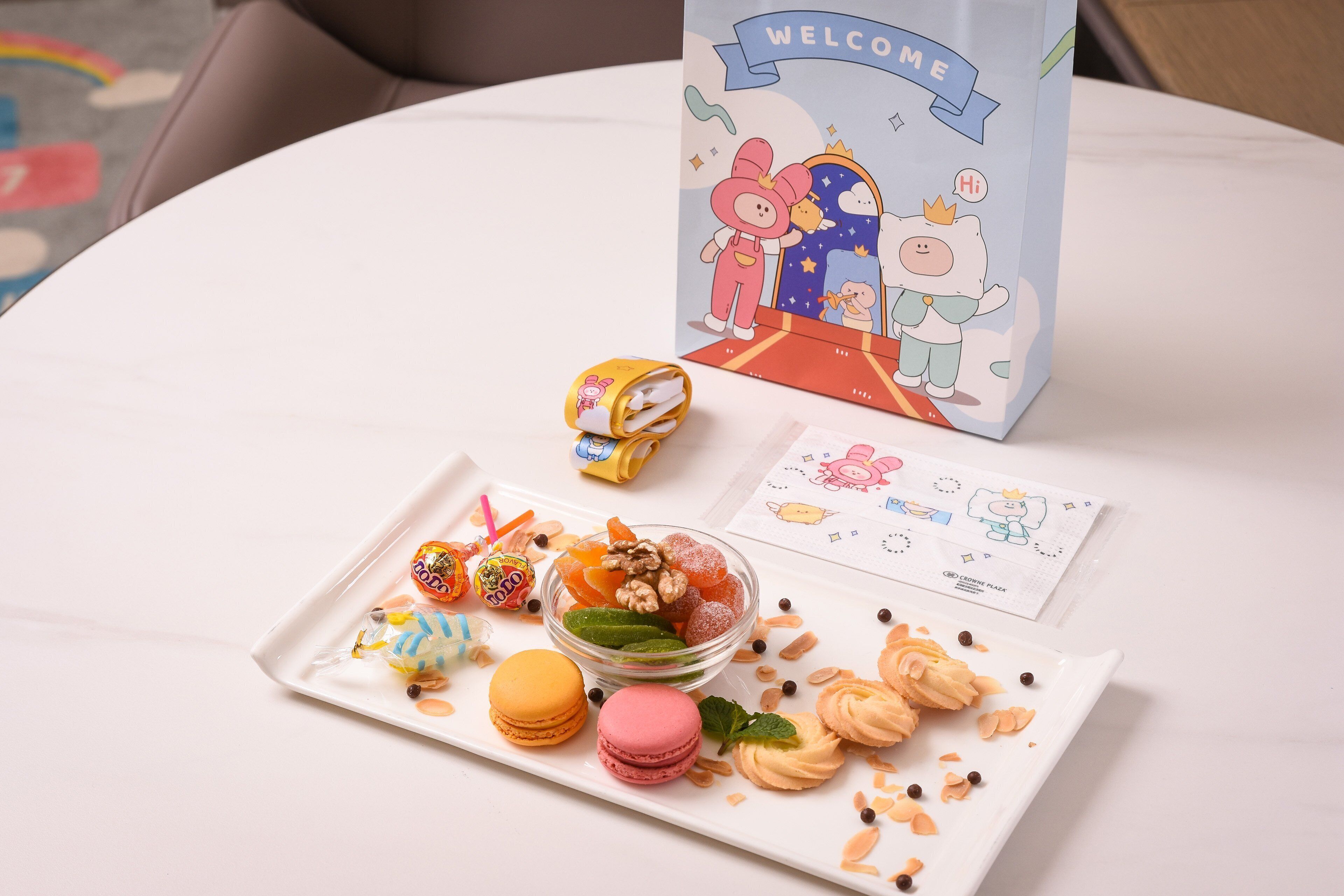 A table with a plate of macarons, a bowl of mixed nuts and dried fruits, and a 'WELCOME' bag.