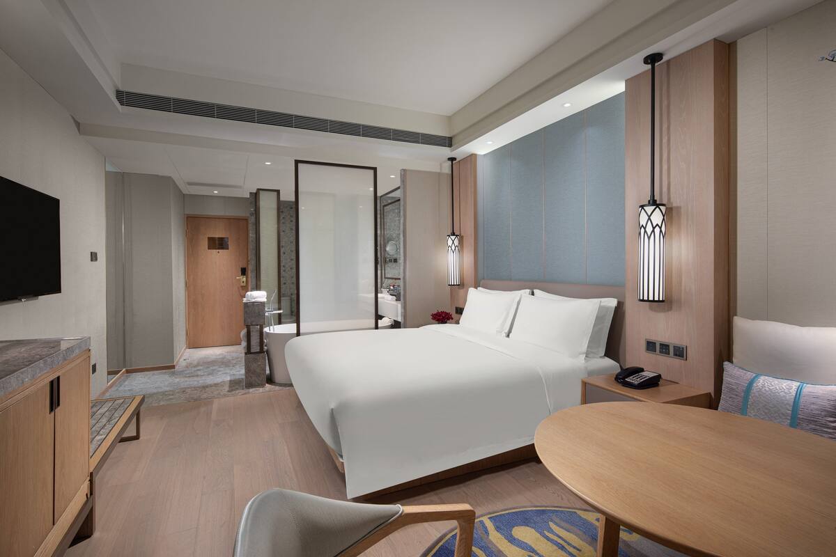 Una habitación de hotel moderna con una cama grande, un escritorio, una silla, un televisor y un baño visible a través de una puerta de cristal.