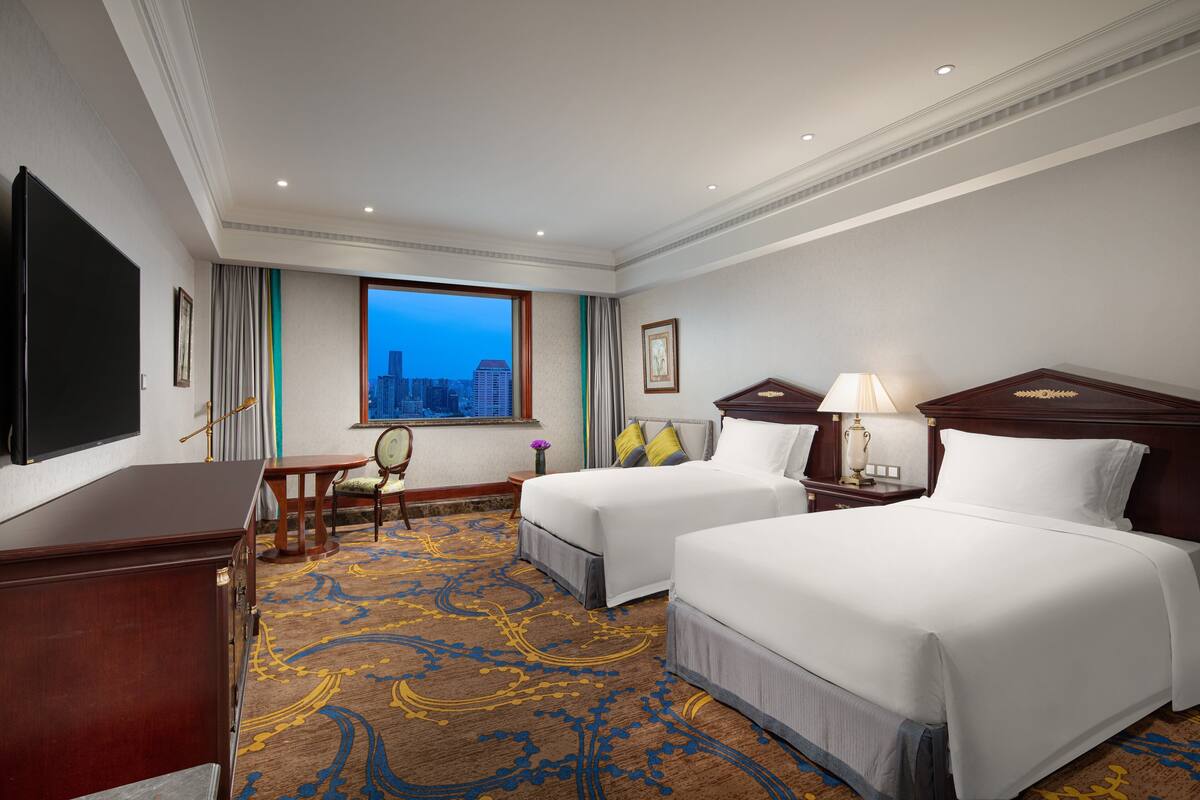 Habitación de hotel con dos camas, un escritorio, una silla, un televisor y una ventana con vista a la ciudad.