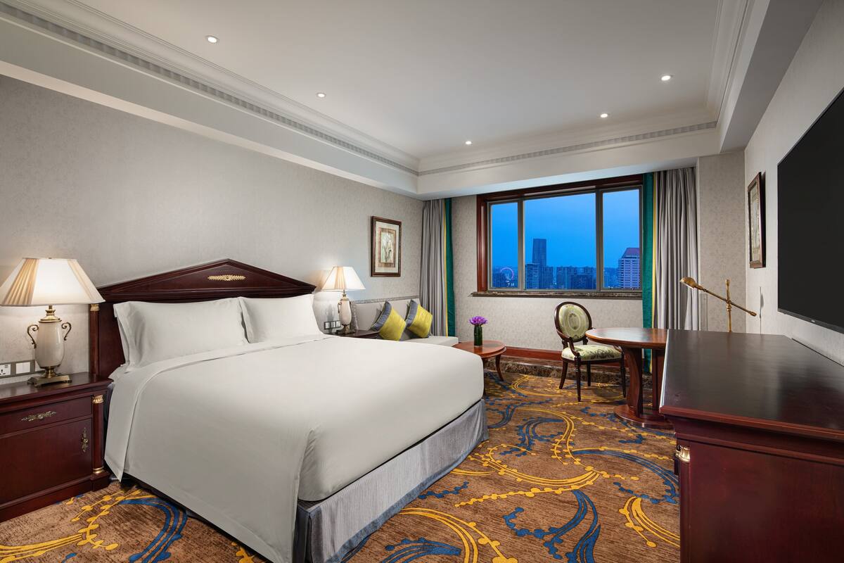 Habitación de hotel con una cama grande, un escritorio, una silla y vistas de la ciudad a través del ventanal.