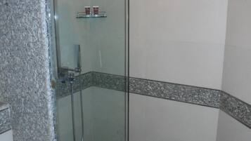 Zona de ducha con paredes de vidrio, cabezal de ducha, ducha de mano y estante de vidrio con artículos de tocador.