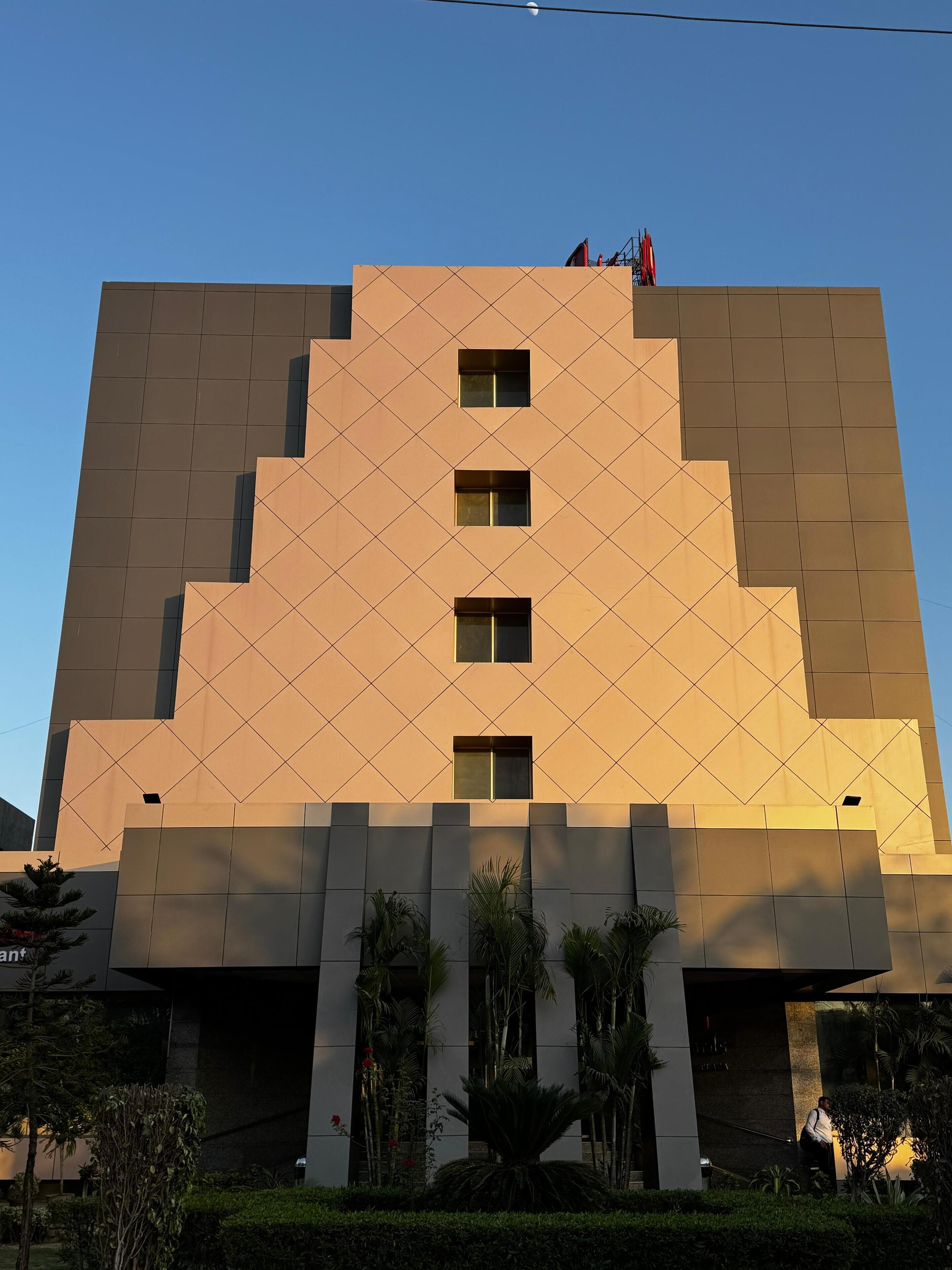 Un edificio moderno con fachada geométrica, múltiples ventanas y un área de entrada ajardinada con palmeras y arbustos.