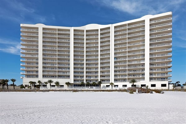 A large, multi-story hotel with numerous balconies, located on a beach with palm trees.