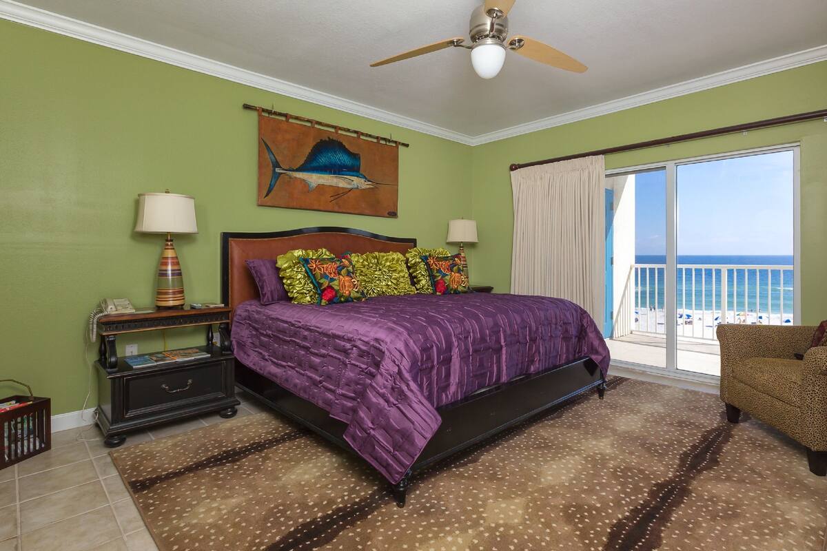 Un dormitorio con cama, ventilador de techo, un cuadro de un pez, mesita de noche con lámpara y vista a la playa por una ventana.