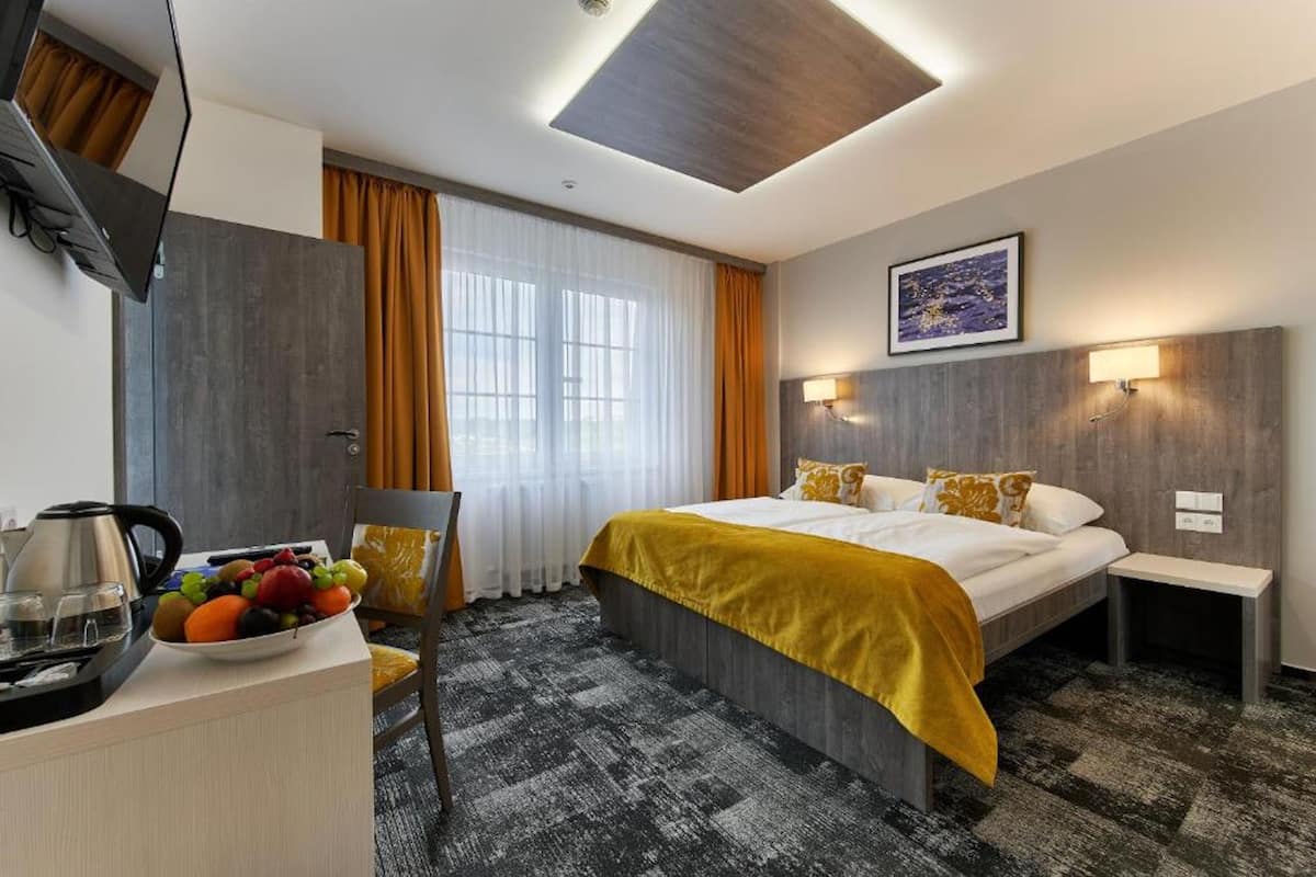 Habitación de hotel con cama, escritorio con hervidor y fruta, televisión y ventana con cortinas.