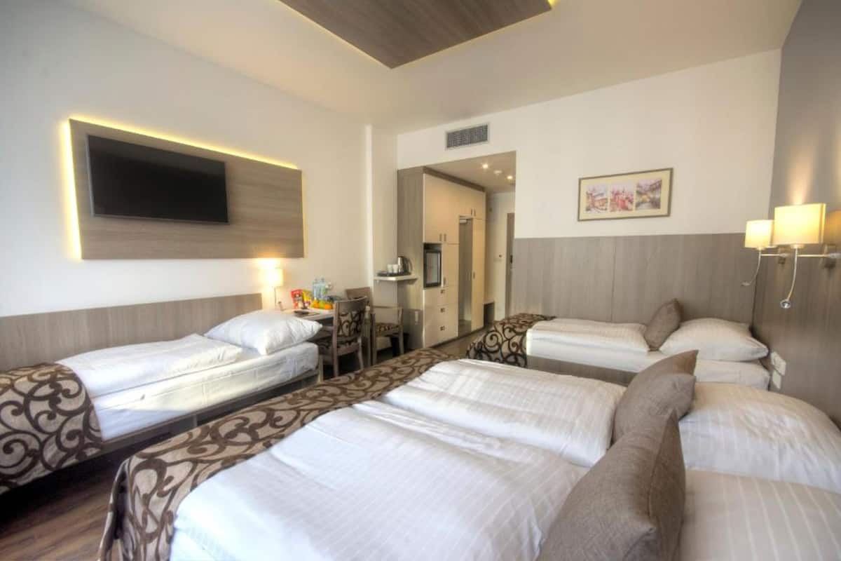 Habitación de hotel con cuatro camas individuales, un escritorio con silla, televisor de pantalla plana montado en la pared y una kitchenette pequeña.