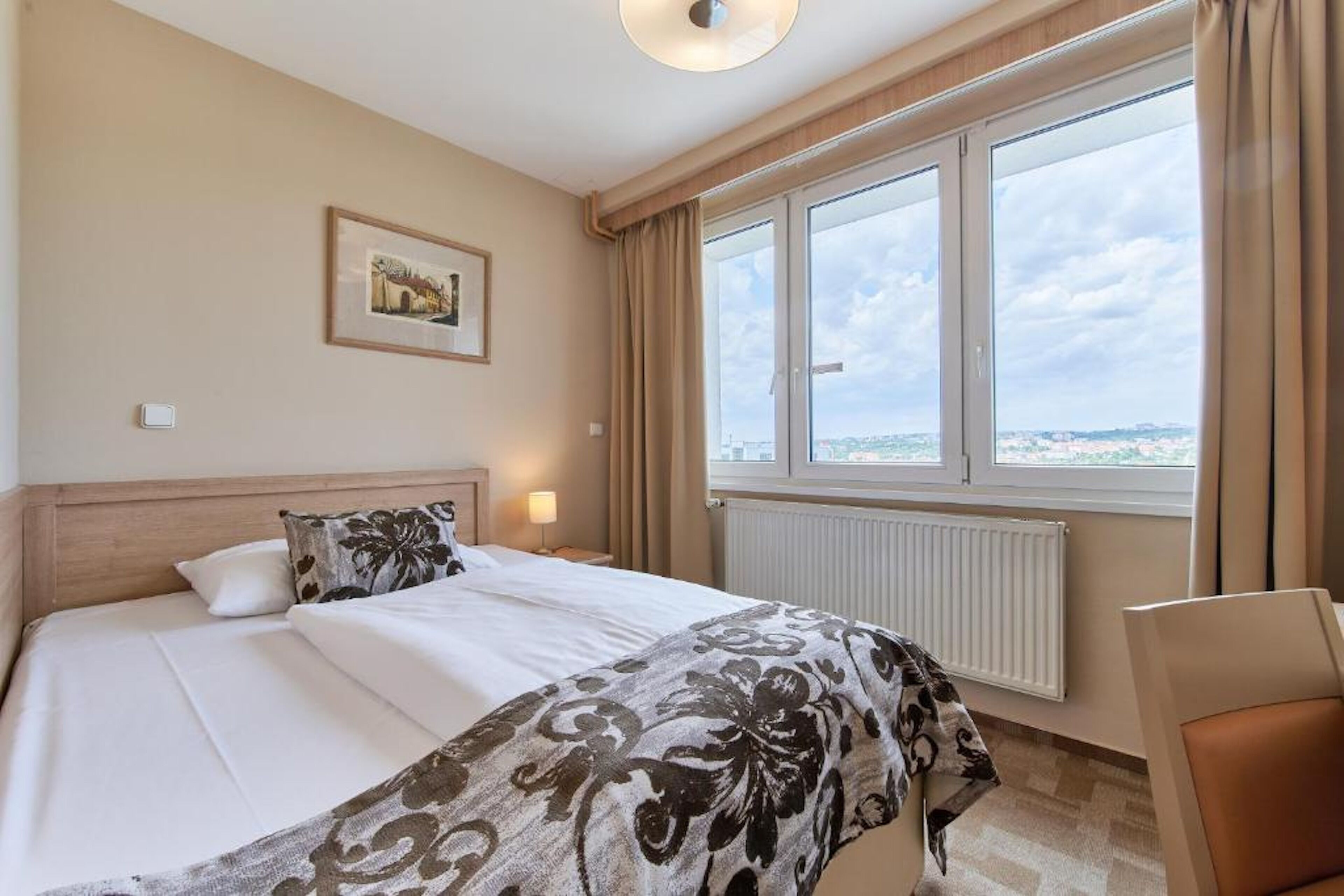 Habitación de hotel con una cama grande, vista de la ciudad a través de una ventana, un cuadro en la pared y una silla junto a la ventana.
