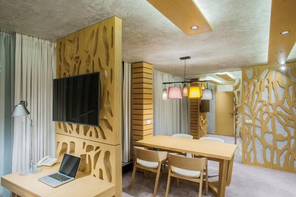 Una habitación de hotel moderna con un separador de madera, un escritorio con una computadora portátil, una zona de comedor con sillas y un televisor montado en la pared.
