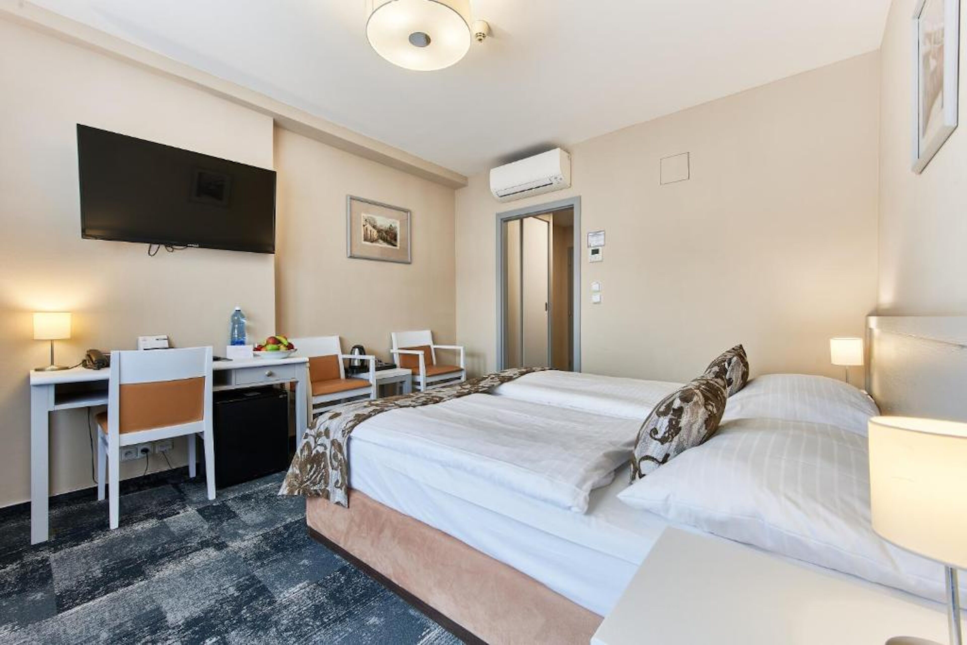 A hotel room with a bed, desk, chairs, a television, and an air conditioning unit.