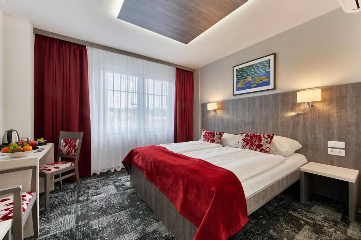Una habitación de hotel con una cama, un escritorio con silla, una cortina roja, una ventana y un cuadro en la pared.