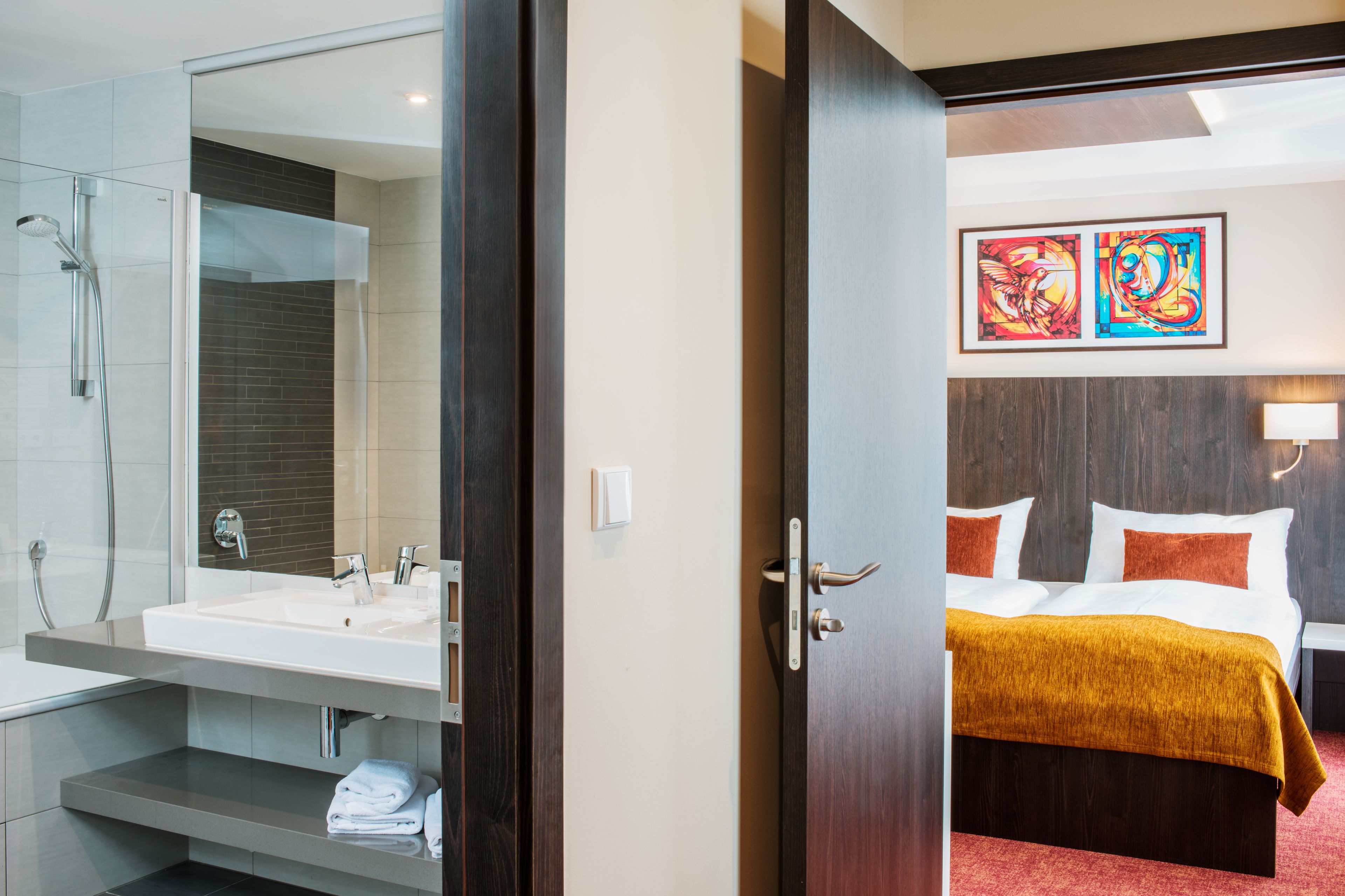 A hotel room with a bathroom featuring a glass-enclosed shower, a sink, and a towel rack. The room has a bed with a brown headboard, white bedding, and a red throw blanket. There are two framed artworks on the wall above the bed.