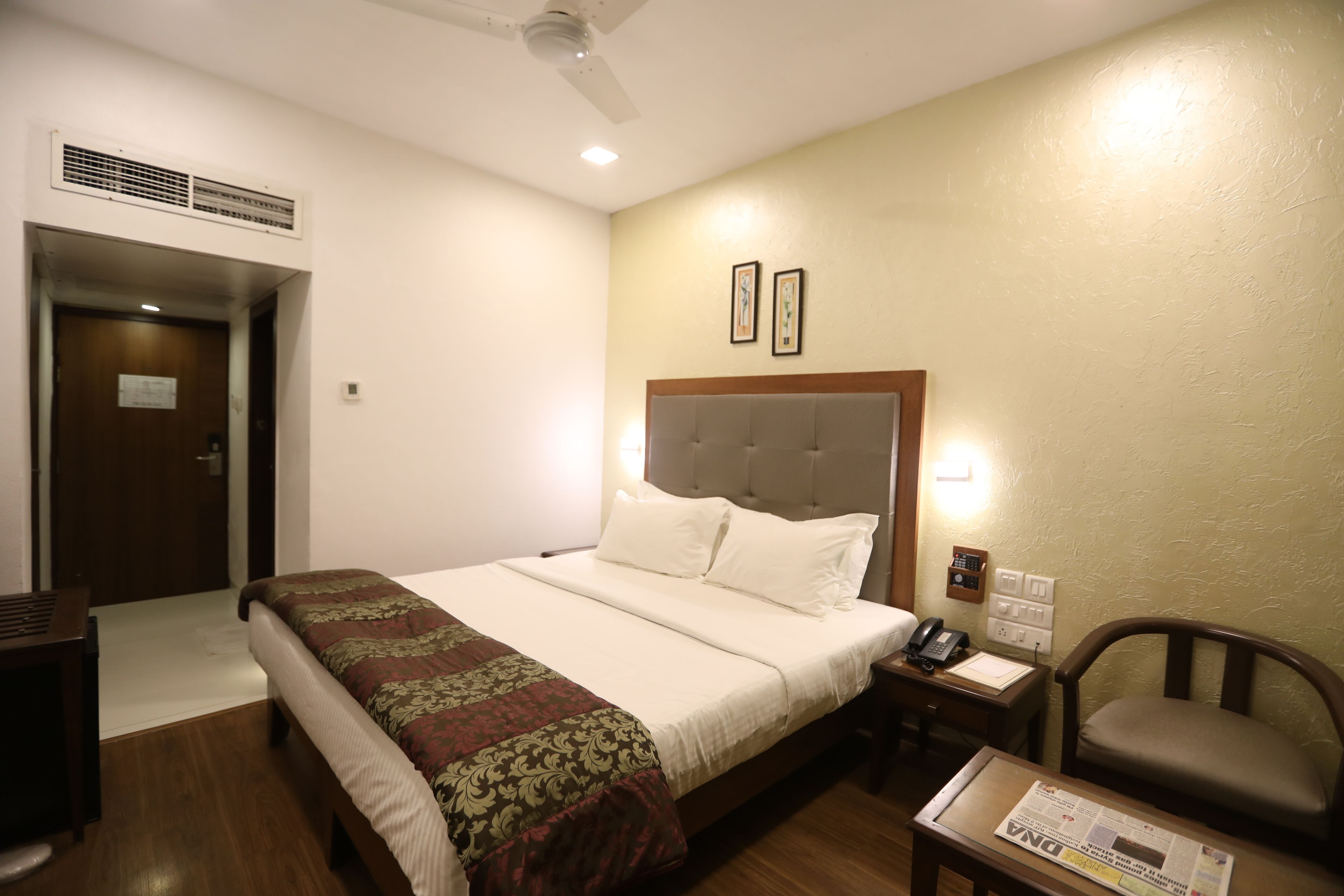A hotel room with a bed, bedside table, chair, and a small table with a newspaper.