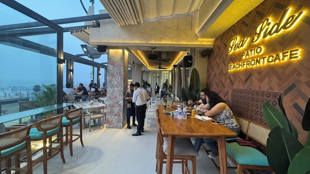A rooftop cafe with a view of the beach, wooden tables and chairs, and a neon sign reading "Side Patio Beachfront Cafe". - Citizen Hotel (Mumbai)