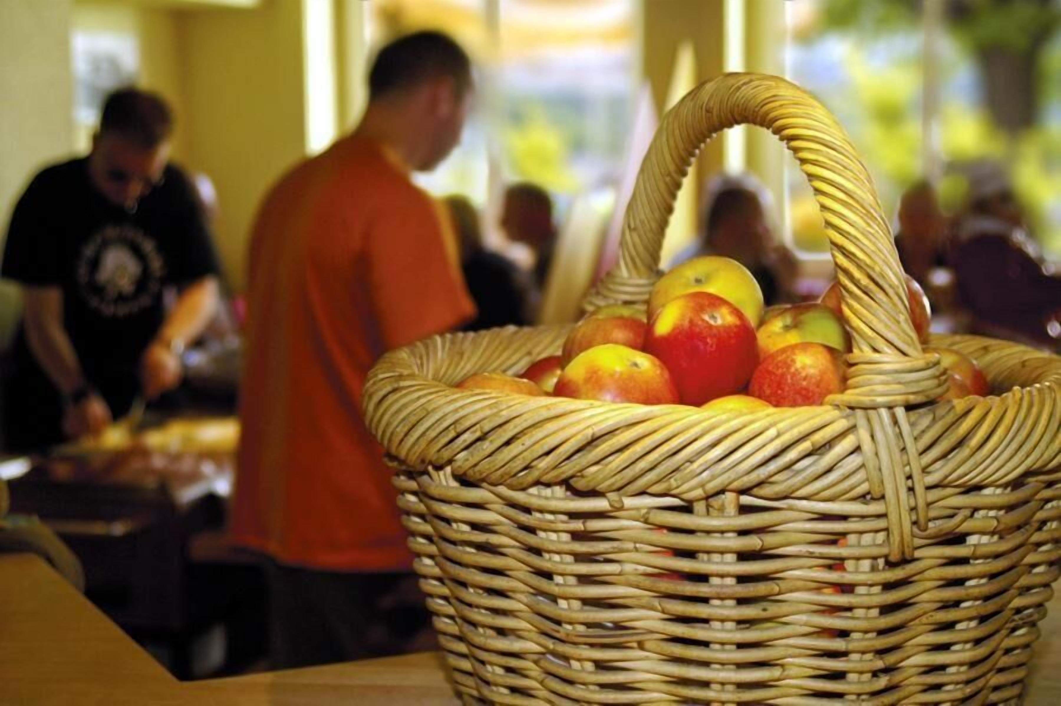A wicker basket filled with apples on a table in a room with people in the background.
