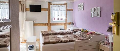 A bedroom with a bed, a TV, a window with curtains, a radiator, and a small table with shoes.
