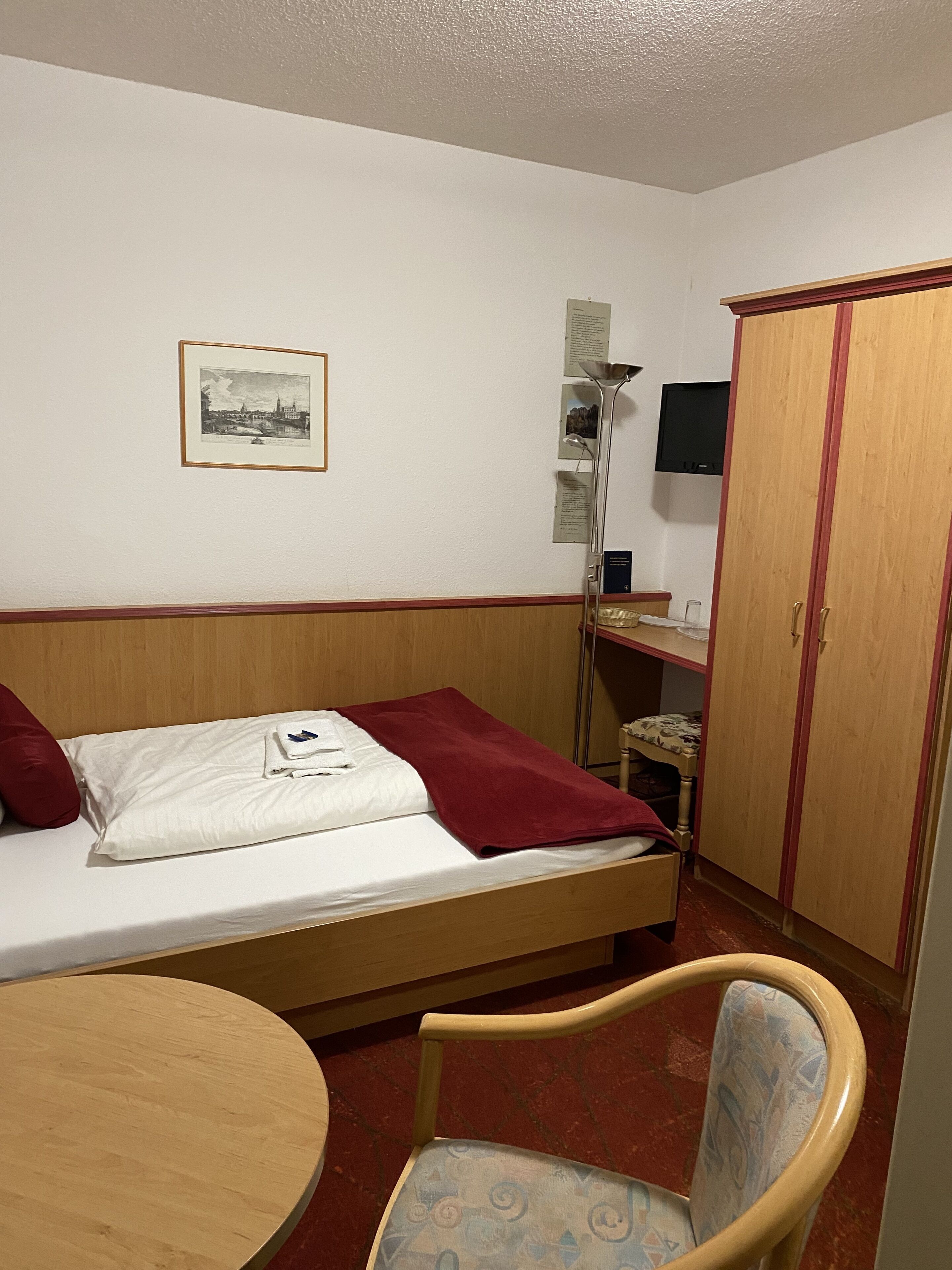 Un lit simple avec une couverture rouge, une tête de lit en bois, une petite table, une chaise, une armoire et un tableau encadré accroché au mur.