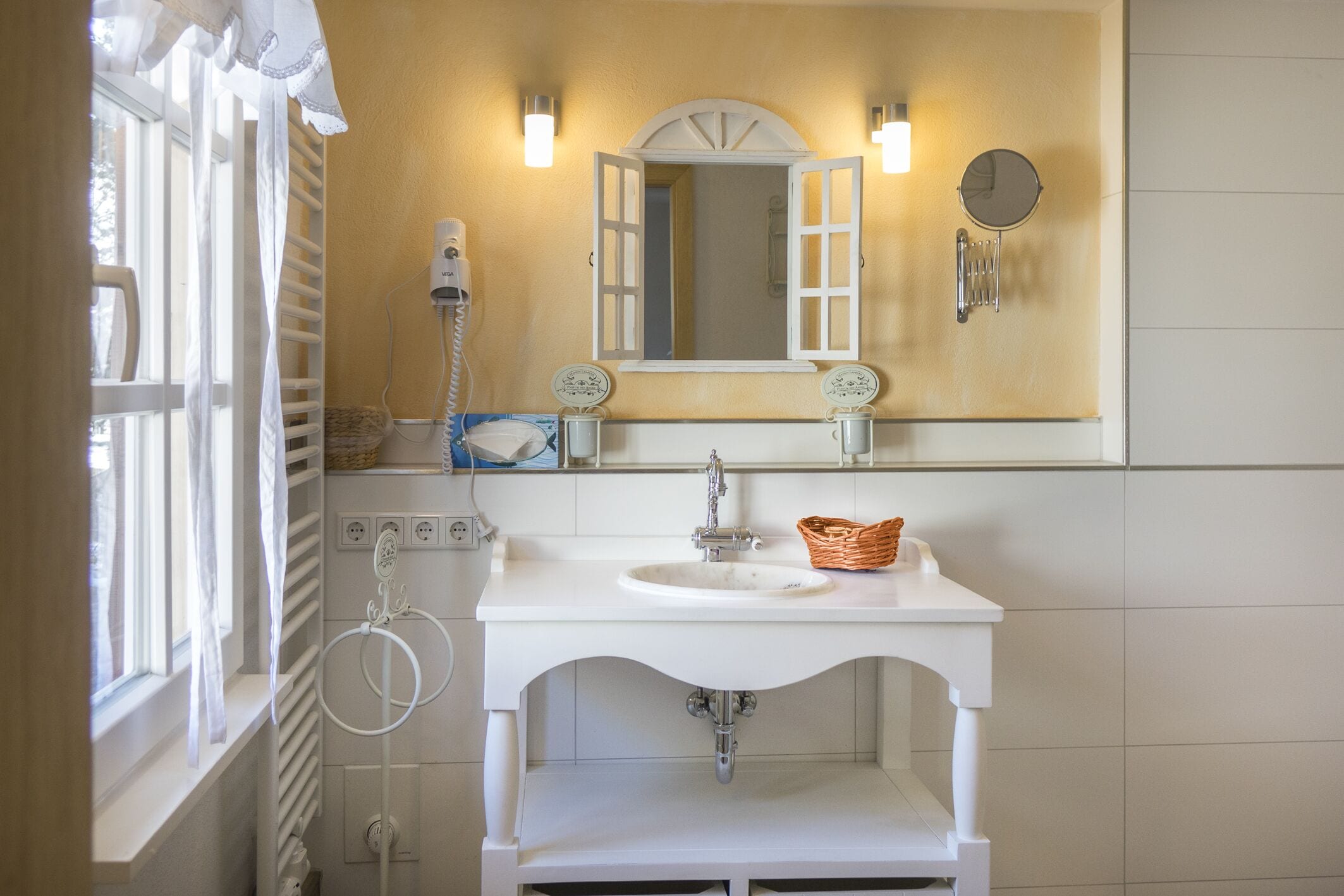 A bathroom with a white sink, a mirror with a white frame, a towel rack, and a window with white curtains.