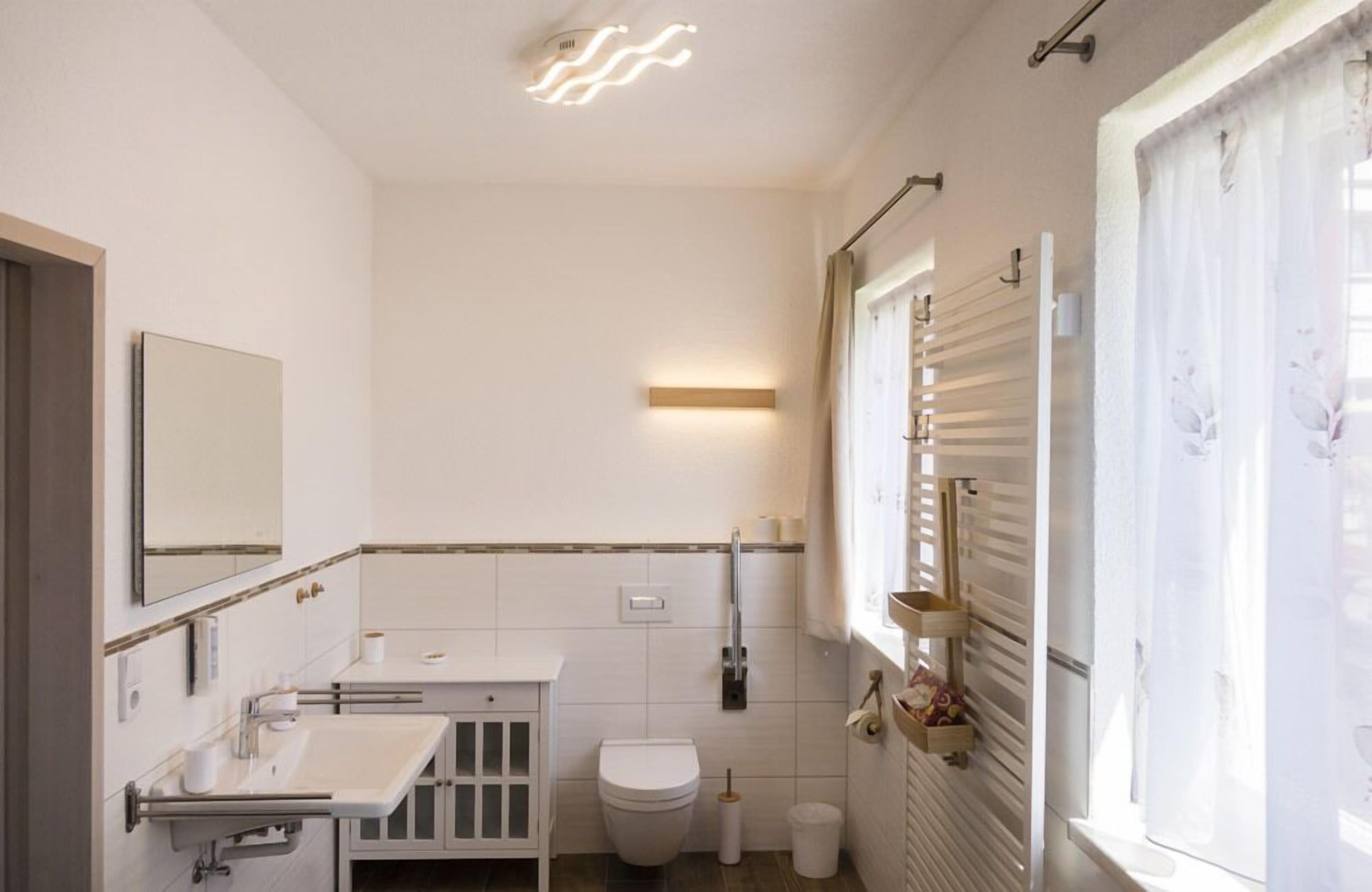 Ein modernes Badezimmer mit Toilette, Waschbecken und Spiegel. Ein Fenster mit Jalousien und ein Handtuchhalter sind vorhanden.