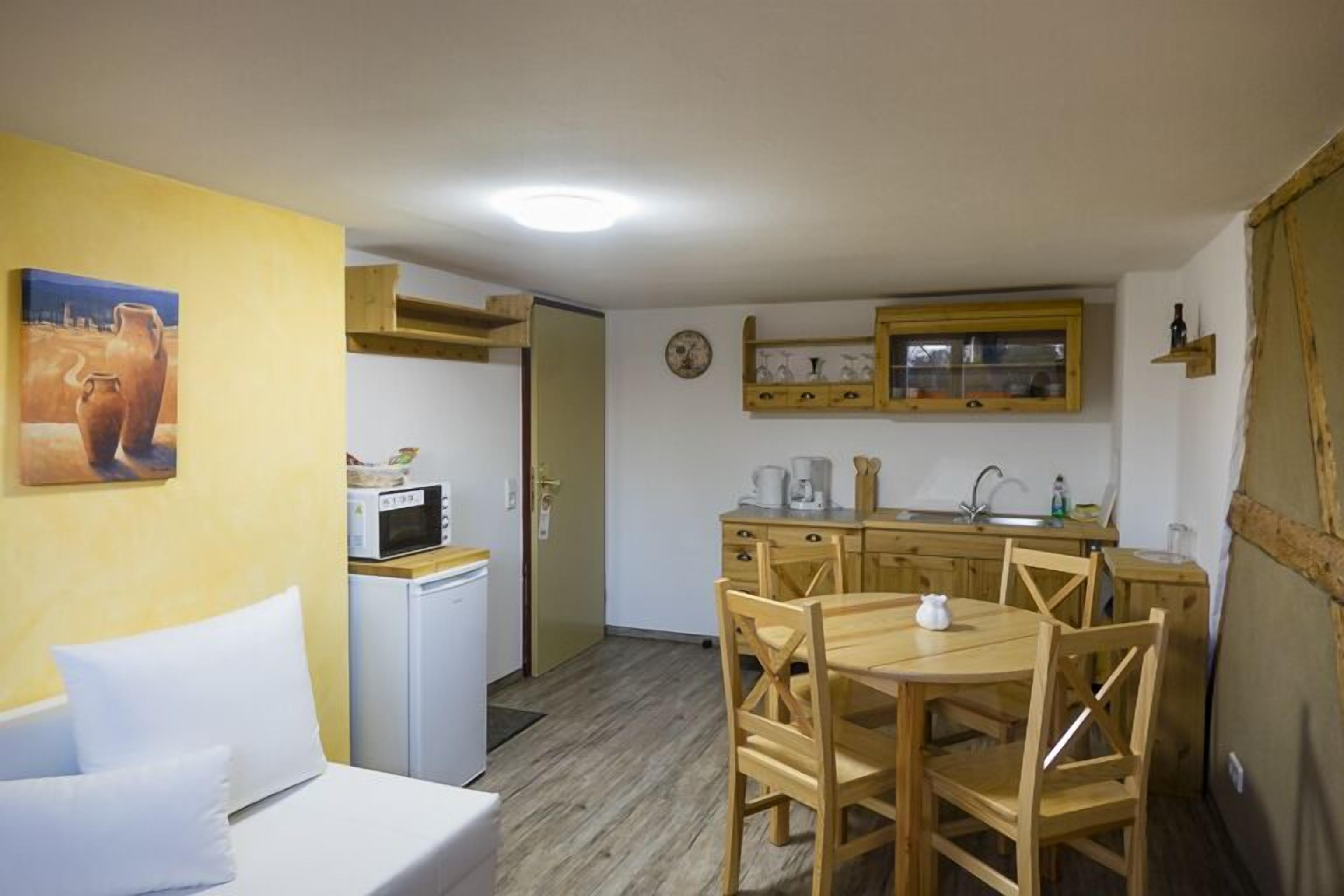 A compact room with a bed, a small kitchenette, a dining area with wooden chairs, and a painting on the wall.