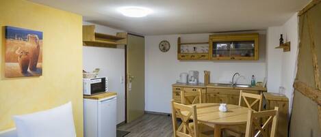 A compact room with a bed, a small kitchenette, a dining area with wooden chairs, and a painting on the wall.