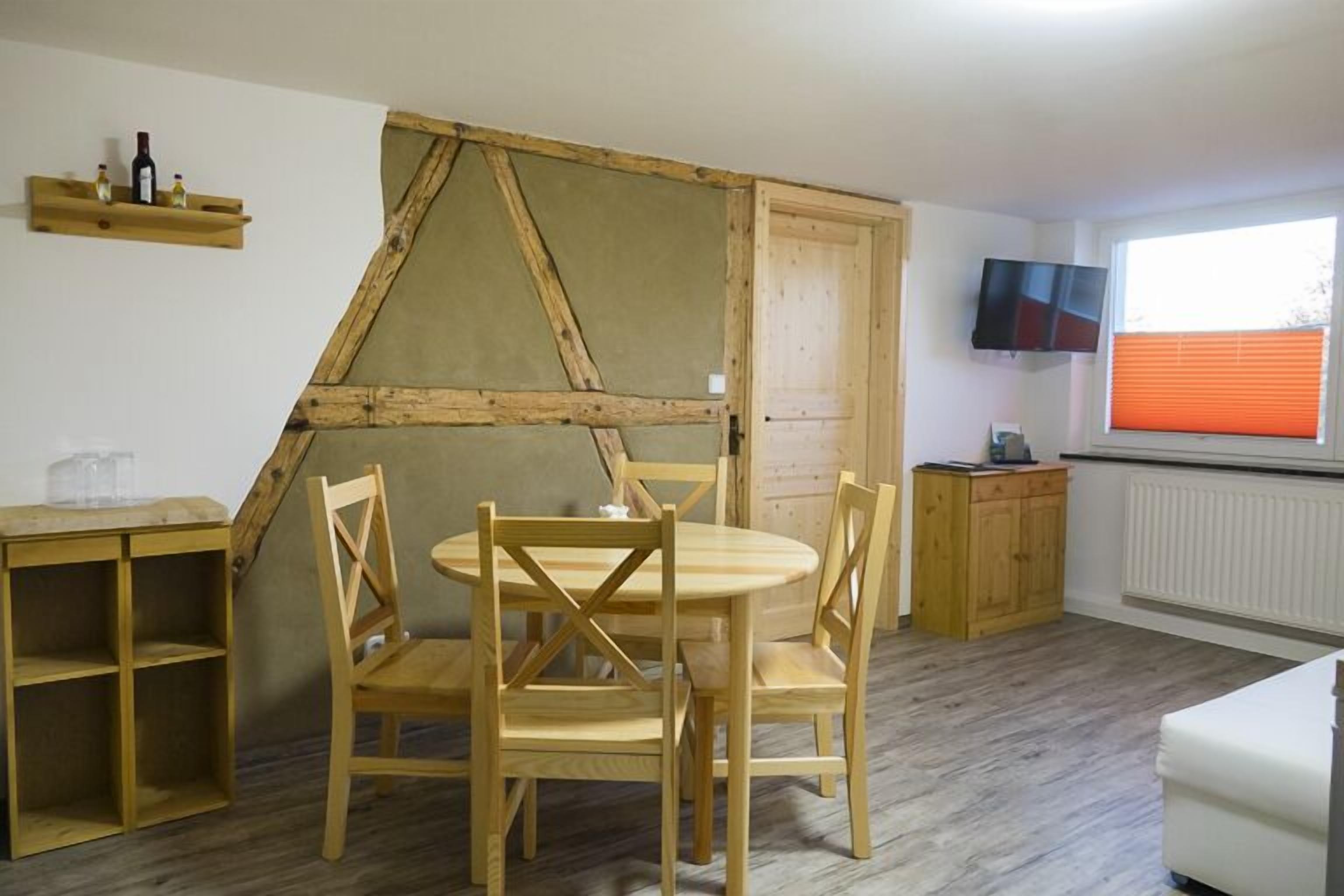 A room with a wooden dining table and chairs, a television on the wall, and a window with blinds.