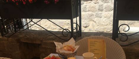 Une table dressée avec des aliments pour le petit-déjeuner, notamment des croissants, des fruits, du fromage et de la charcuterie, avec une chaise en osier et un mur de pierre en arrière-plan.