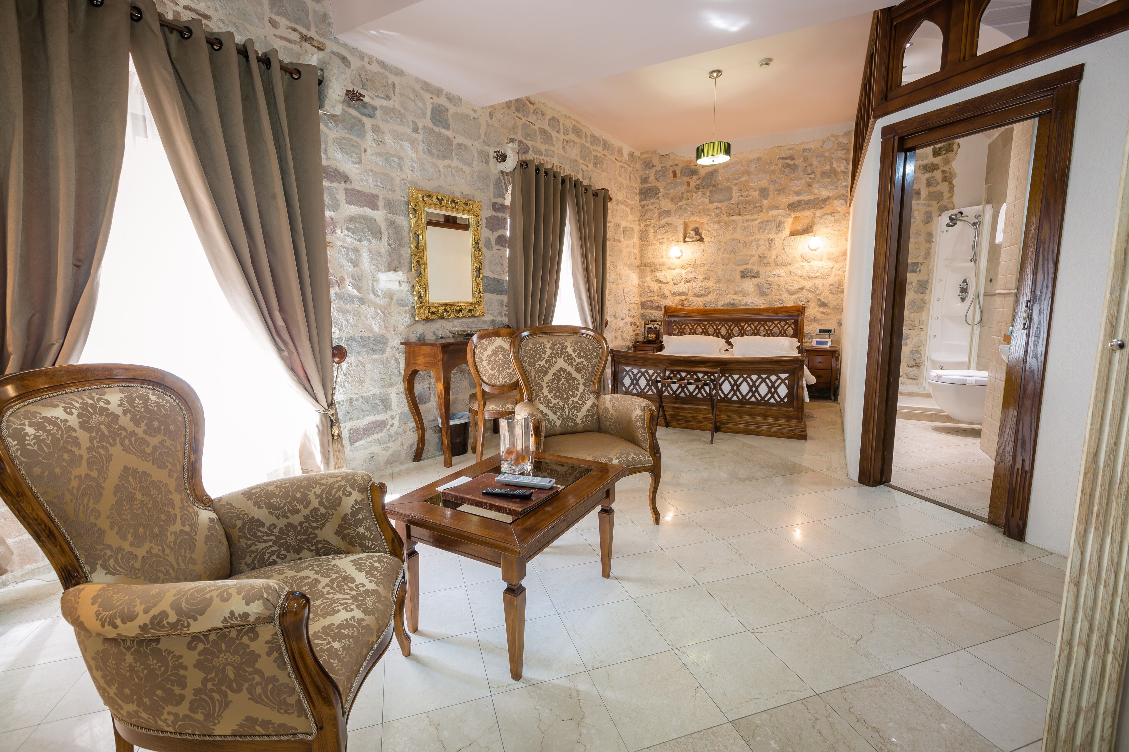 A room with a stone wall, a wooden bed, a wooden coffee table with books, and two patterned armchairs.