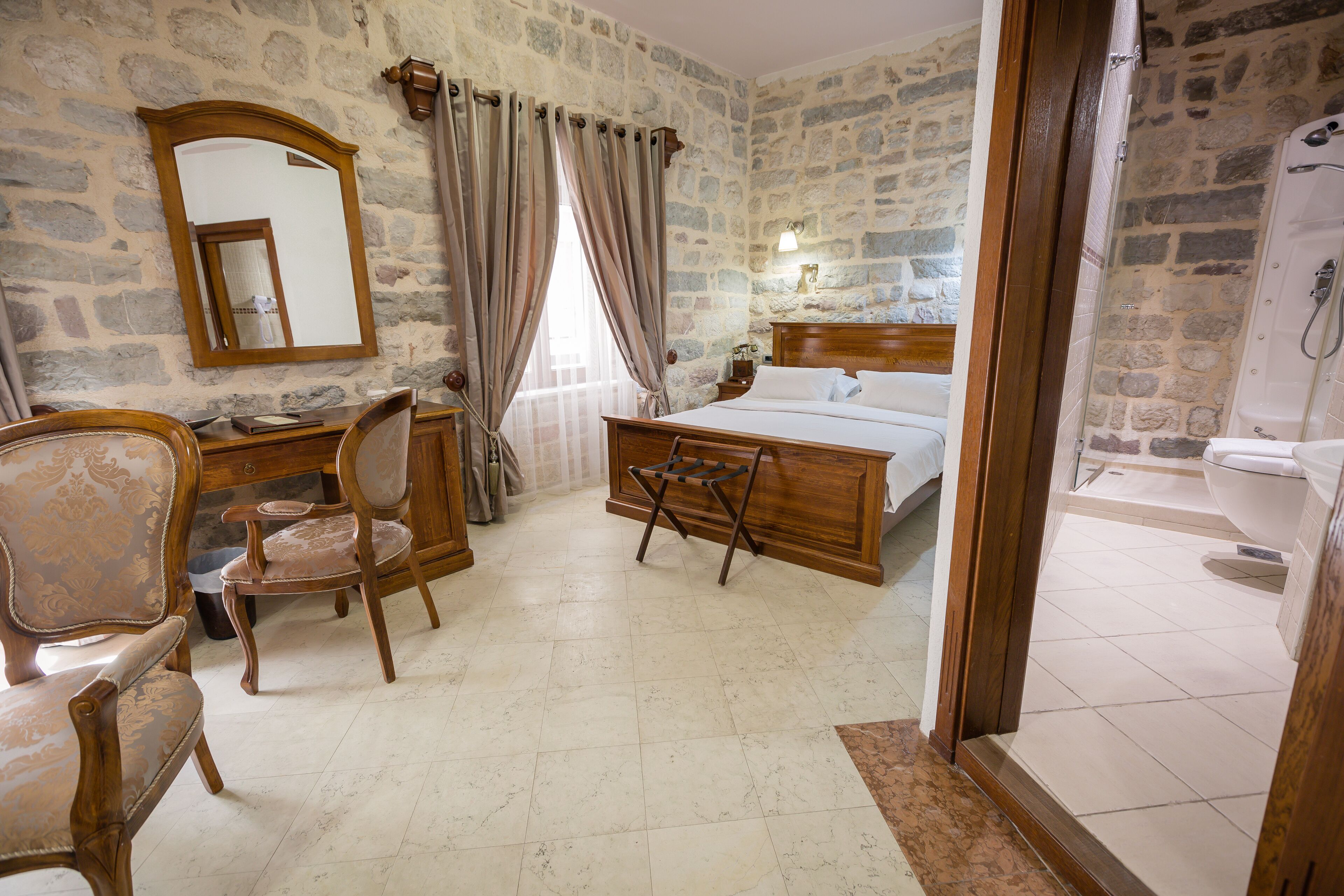 A room with a stone wall, a wooden bed, a desk with a chair, and a bathroom with a bathtub.