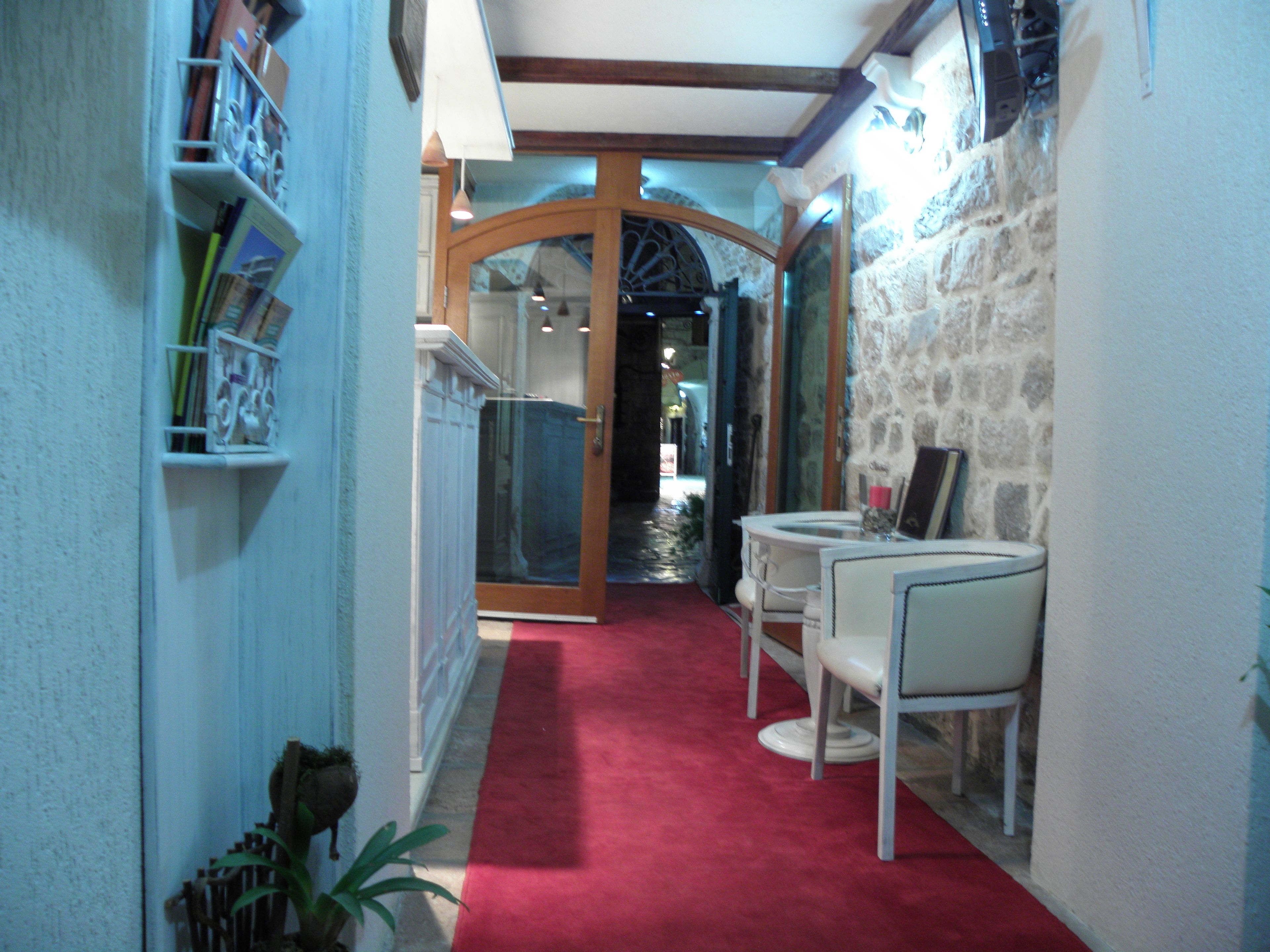 A narrow hallway with a red carpet, white walls, and a stone wall on one side. There is a white table with chairs and a small plant on the left.