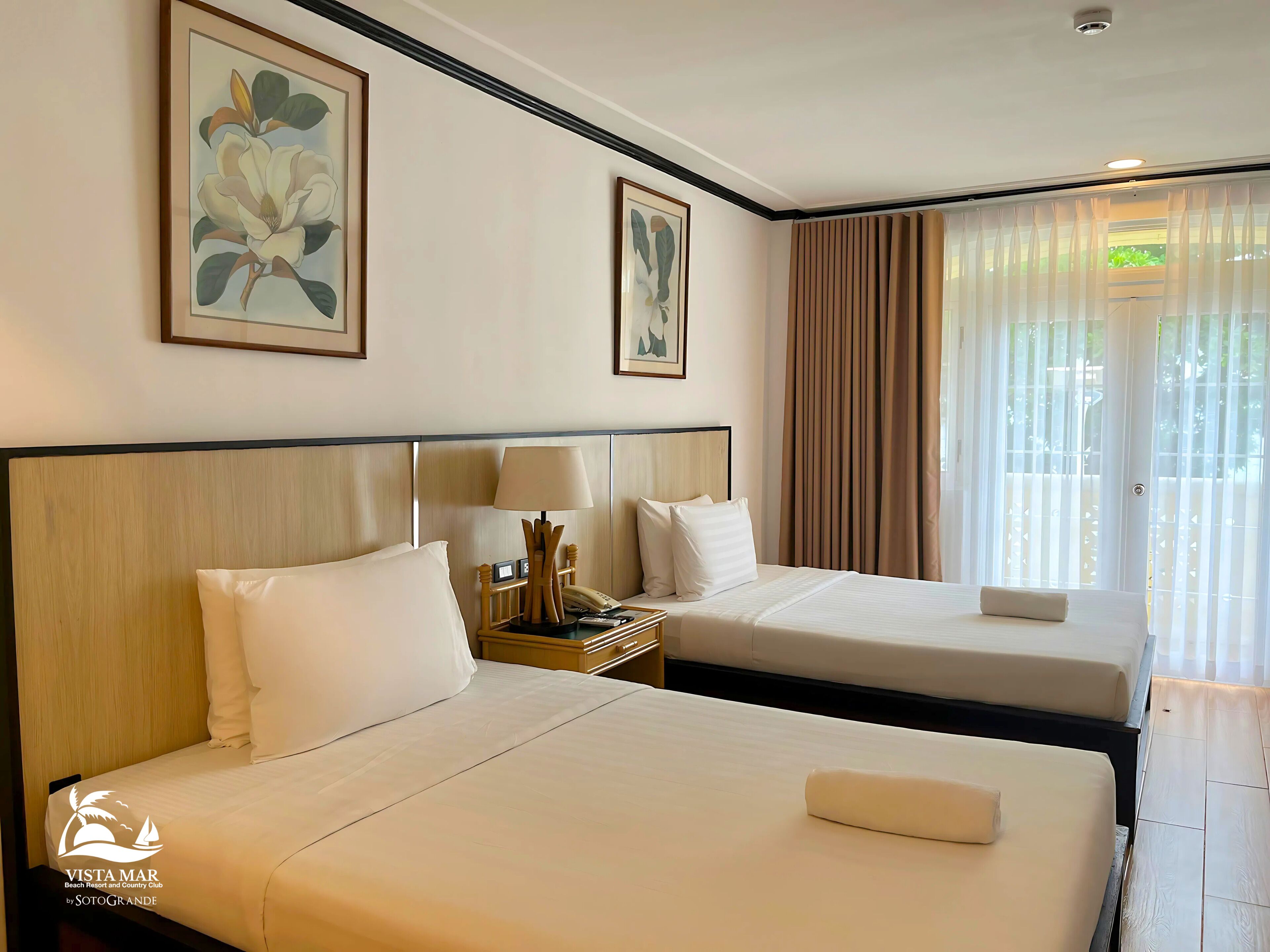 A hotel room with two beds, a nightstand with a lamp, and artwork on the walls.
