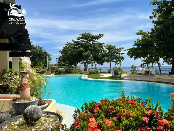 A clear blue pool surrounded by tropical landscaping, with a view of the ocean and a beach resort in the background.