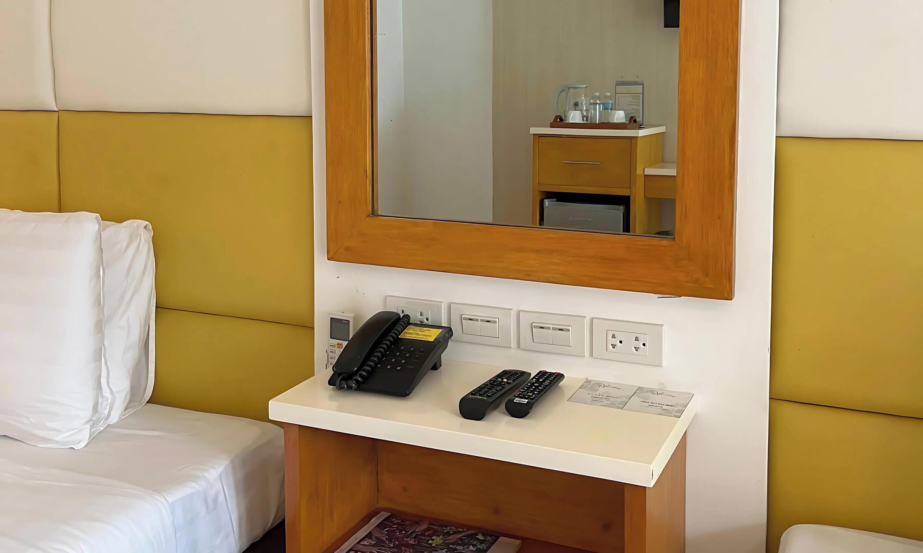 A hotel room with a bed, a bedside table with a phone and remote controls, a mirror, and a wall socket.