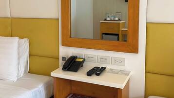 A hotel room with a bed, a bedside table with a phone and remote controls, a mirror, and a wall socket.