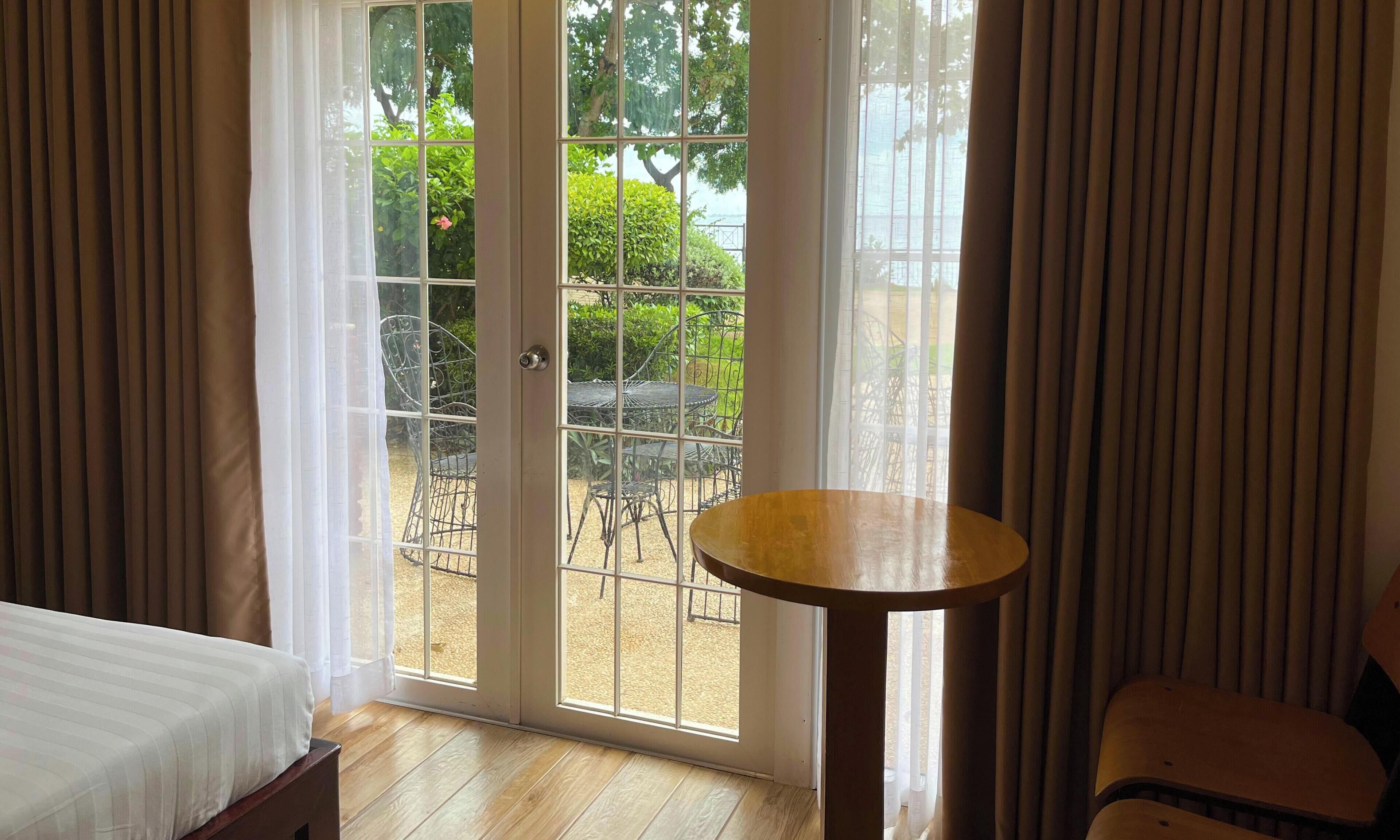 Une chambre d’hôtel avec un lit, une petite table ronde et une porte-fenêtre coulissante donnant sur une terrasse extérieure avec une table et des chaises.