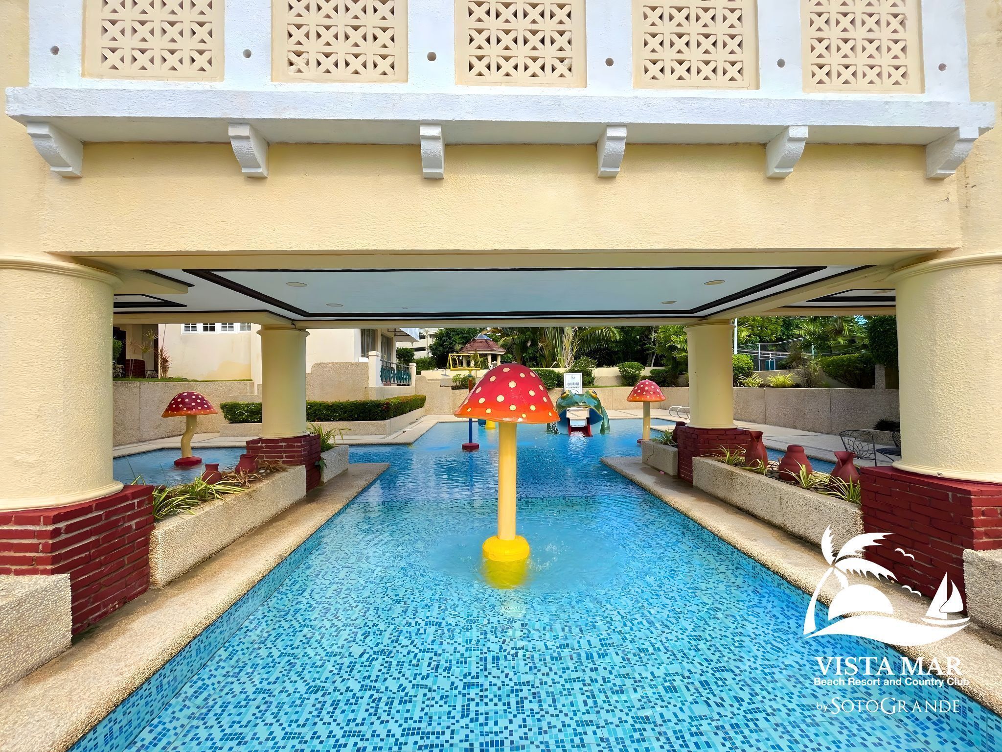 Una piscina con esculturas decorativas de hongos, un paseo techado y vistas de la arquitectura del complejo.