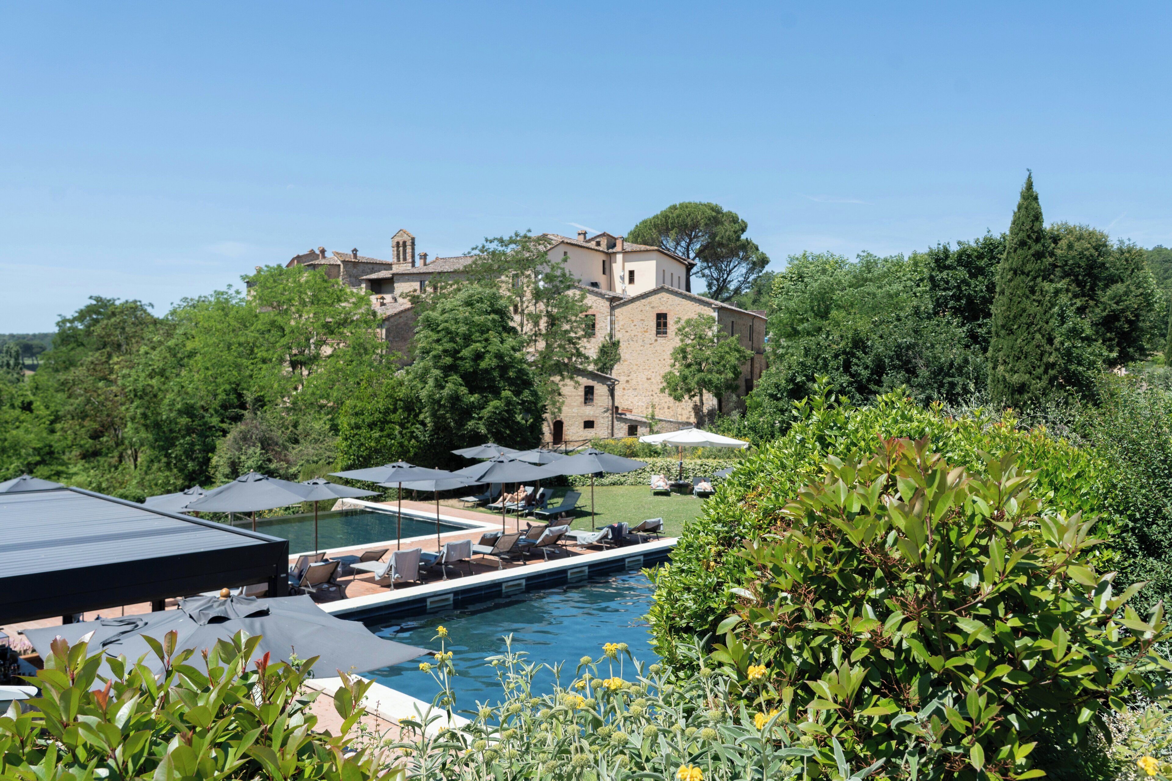 A pool area with lounge chairs and umbrellas, surrounded by greenery and a historic building in the background.